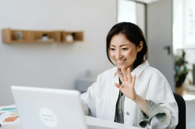 Smiling woman waves at laptop during a video call in a modern office.